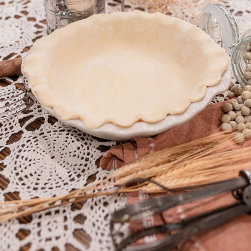 An unbaked sourdough pie crust fitted into a white ceramic pie dish with a crimped ruffled edge, surrounded by a vintage flour sifter, a glass jar of dried beans for blind baking, dried grain stalks, and vintage pastry tools on a crocheted doily over dark wood.