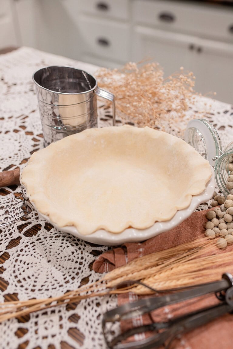 An unbaked sourdough pie crust fitted into a white ceramic pie dish with a crimped ruffled edge, surrounded by a vintage flour sifter, a glass jar of dried beans for blind baking, dried grain stalks, and vintage pastry tools on a crocheted doily over dark wood.