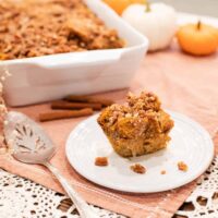 A slice of sourdough pumpkin French toast casserole on a plate with cinnamon sticks and a casserole dish in the background, accompanied by autumn decor.