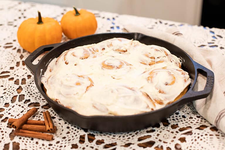 A cast iron skillet filled with frosted sourdough pumpkin spice cinnamon rolls, surrounded by pumpkins and cinnamon sticks, ready to be served at a cozy fall gathering.