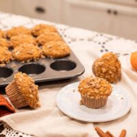 A display of sourdough pumpkin spice muffins, with a couple placed on a plate, showcasing their golden color and streusel topping, surrounded by mini pumpkins and a muffin tin full of more muffins.