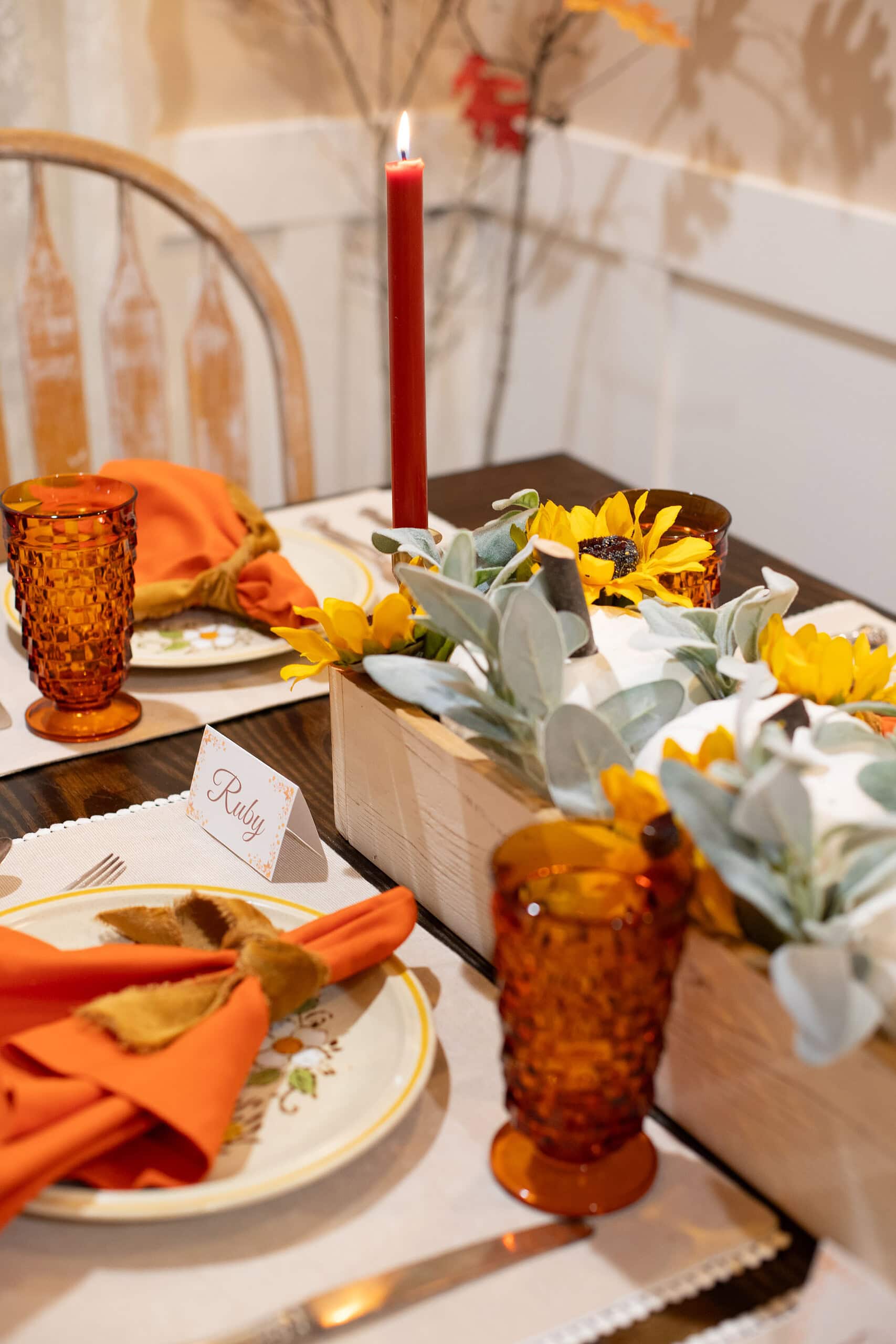 Angled close-up of a cottage fall tablescape showing a place card reading "Ruby" beside an orange napkin with velvet tie on a vintage floral plate, with the wood box centerpiece of white pumpkins, sunflowers, and lamb's ear, amber hobnail goblets, and a lit red taper candle in a brass candlestick.