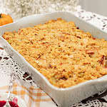 A close-up of the baked sourdough cornbread dressing in a ceramic baking dish, with a spoon ready to serve. The dish’s golden-brown texture captures the rustic appeal of a comforting holiday side dish.