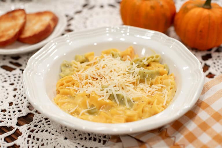 A serving of creamy pumpkin mac and cheese in a white bowl, topped with shredded cheese. The colorful autumn-themed pasta is coated in a thick, orange pumpkin sauce, with toasted bread slices and small pumpkins in the background.