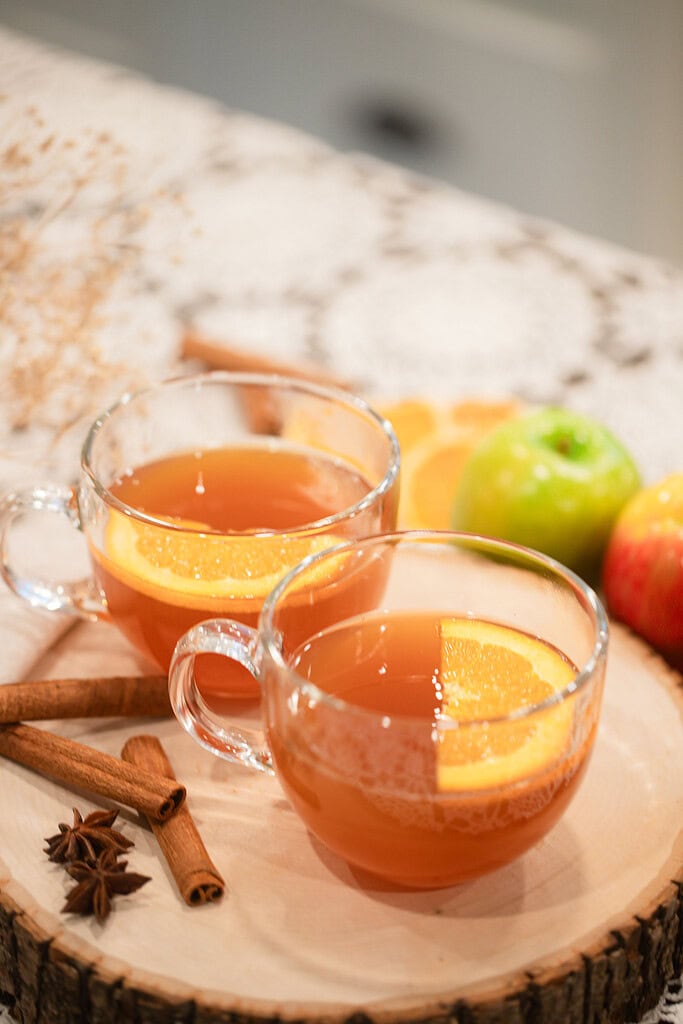 Two glass mugs of warm spiced apple cider garnished with orange slices on a wood slice serving board, styled with cinnamon sticks, star anise, and green and red apples on a lace doily.