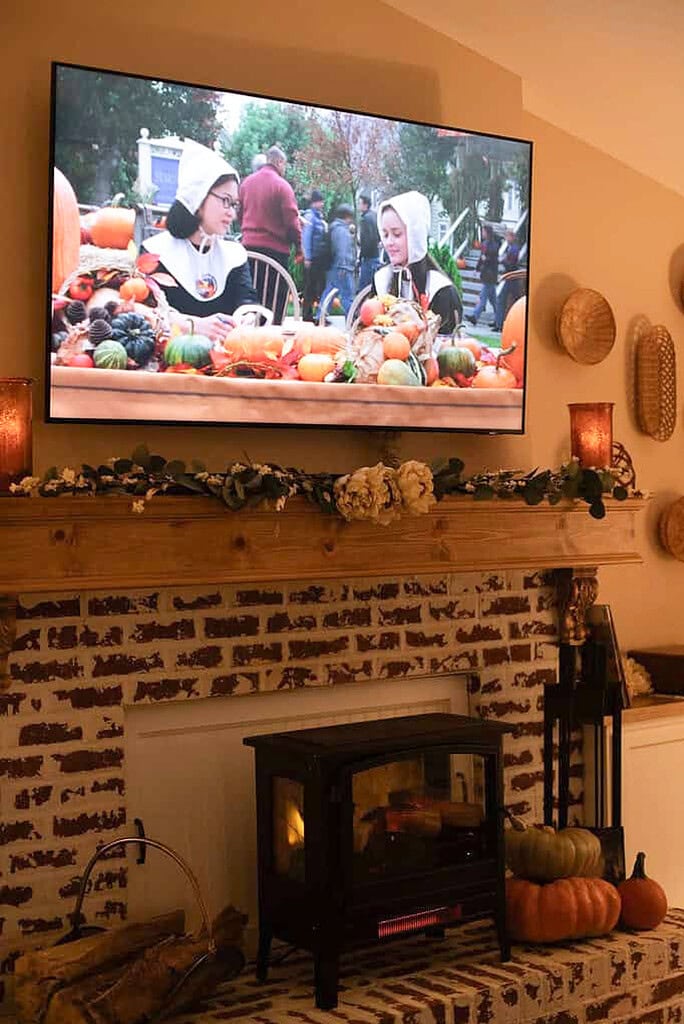 A cozy cottage living room with a fall-decorated brick fireplace and wood mantel adorned with a greenery garland and dried flowers, an electric stove insert glowing warmly, pumpkins on the hearth, and a fall movie playing on the wall-mounted television above.