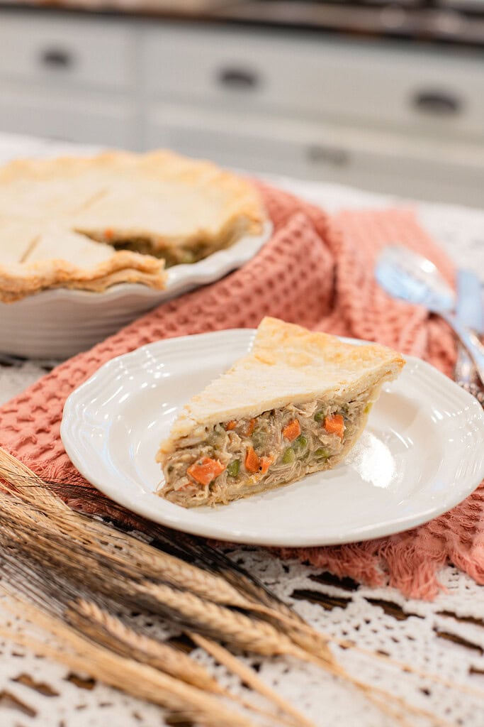 A slice of homemade chicken pot pie with a golden flaky crust revealing shredded chicken, carrots, and peas on a white plate, with the whole pie in a ceramic dish softly blurred on a dusty rose waffle linen behind it.
