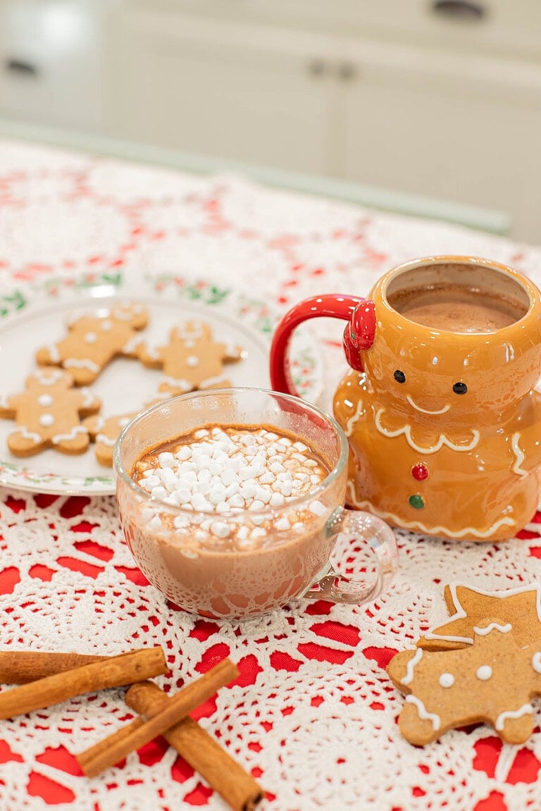 gingerbread hot chocolate with marshmallows in glass mug with gingerbread cookies and festive mug on lace tablecloth