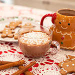Wide shot of a cozy holiday scene featuring gingerbread hot chocolate served in a clear glass mug and a gingerbread mug, surrounded by cinnamon sticks and festive gingerbread cookies on a red and white lace tablecloth.