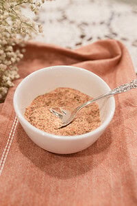 A small white ramekin filled with homemade poultry seasoning blend and a silver vintage spoon, styled on a dusty rose linen with dried baby's breath and a lace doily in the background.