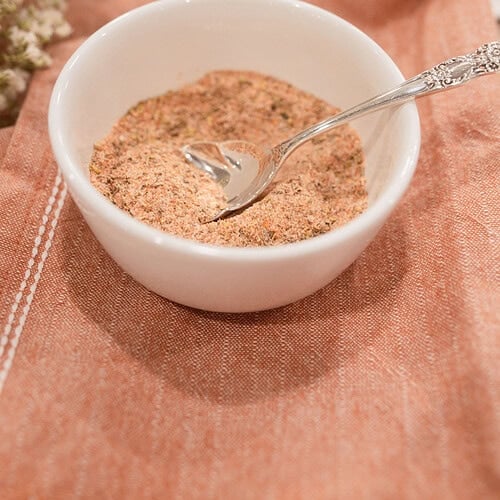 A small white ramekin filled with homemade poultry seasoning blend and a silver vintage spoon, styled on a dusty rose linen with dried baby's breath and a lace doily in the background.