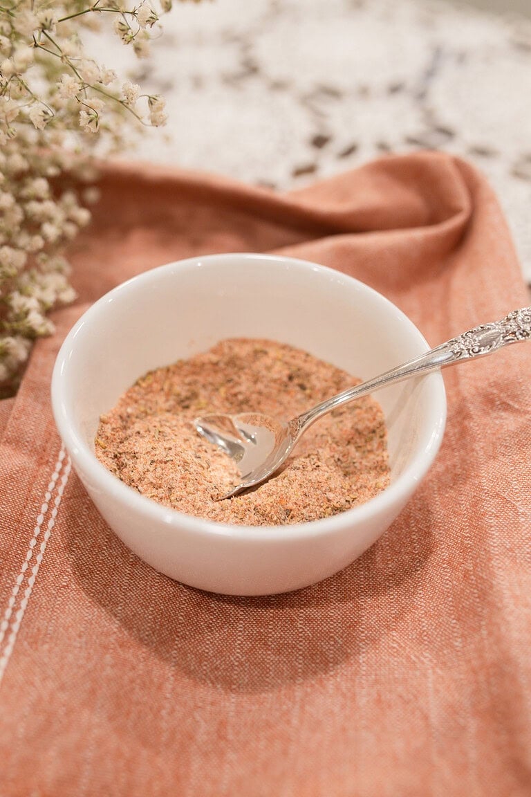 A small white ramekin filled with homemade poultry seasoning blend and a silver vintage spoon, styled on a dusty rose linen with dried baby's breath and a lace doily in the background.