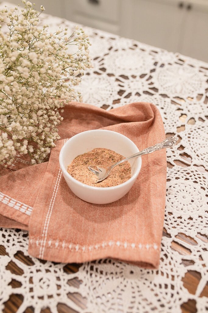 A small white ramekin of homemade poultry seasoning with a silver spoon resting on a folded dusty rose linen, surrounded by dried baby's breath on a lace doily in a cottage kitchen.