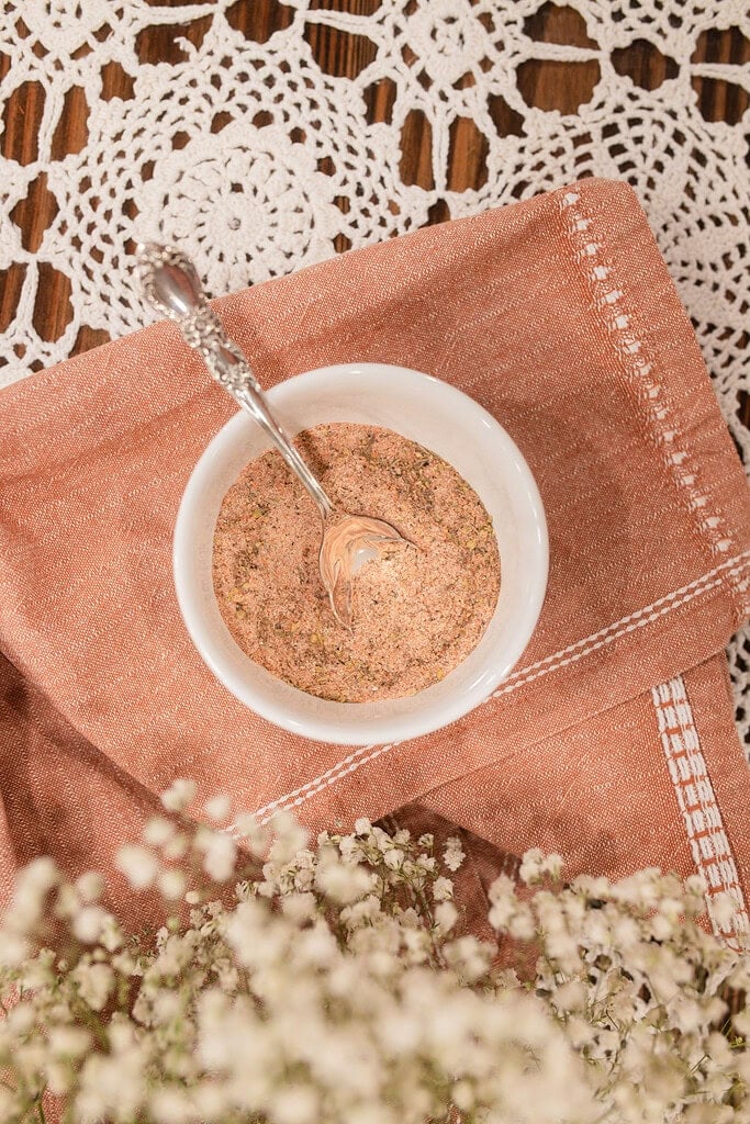 Overhead view of a small white ramekin filled with homemade poultry seasoning and a silver spoon, placed on a folded dusty rose linen on a lace doily with dried baby's breath in the foreground.