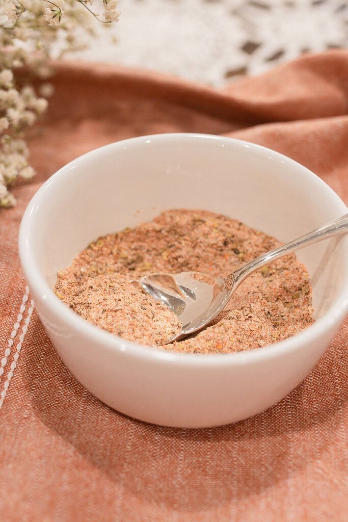 Close-up of a white ramekin filled with homemade poultry seasoning blend and a silver vintage spoon, on a dusty rose linen with dried baby's breath softly blurred in the background.