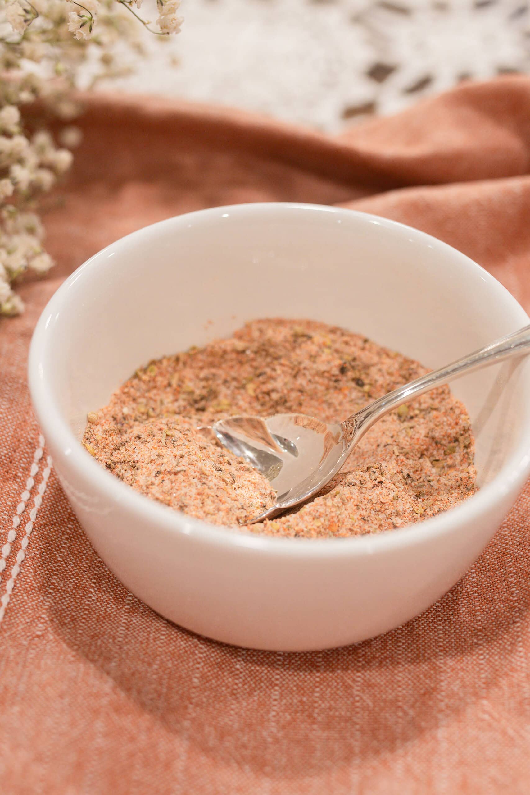 Close-up of a white ramekin filled with homemade poultry seasoning blend and a silver vintage spoon, on a dusty rose linen with dried baby's breath softly blurred in the background.