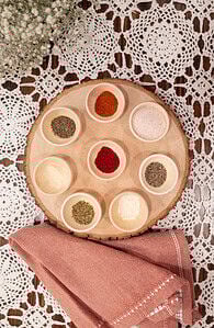 A rustic wooden tray with small bowls filled with different spices and herbs, arranged in a circular pattern. The tray sits on a lace tablecloth, with a pink cloth napkin and delicate baby’s breath flowers nearby, creating a cozy and inviting presentation.