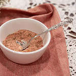 A bowl of poultry seasoning with a silver spoon, set on a pink napkin and lace tablecloth. The surrounding baby’s breath flowers and soft lighting create a warm, cottage-inspired presentation.