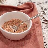 A bowl of poultry seasoning with a silver spoon, set on a pink napkin and lace tablecloth. The surrounding baby’s breath flowers and soft lighting create a warm, cottage-inspired presentation.
