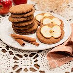 A stack of sourdough ginger apple snap cookies alongside two cookies topped with dried apple slices on a white plate, styled with cinnamon sticks, red apples, and dried botanicals on a lace doily in a cottage kitchen.