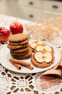 A stack of sourdough ginger apple snap cookies alongside two cookies topped with dried apple slices on a white plate, styled with cinnamon sticks, red apples, and dried botanicals on a lace doily in a cottage kitchen.