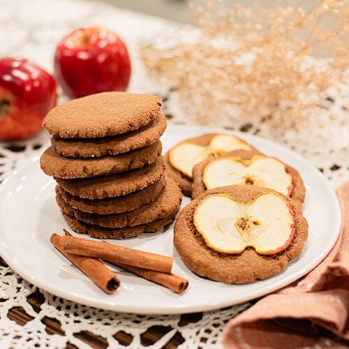 A stack of sourdough ginger apple snap cookies alongside two cookies topped with dried apple slices on a white plate, styled with cinnamon sticks, red apples, and dried botanicals on a lace doily in a cottage kitchen.