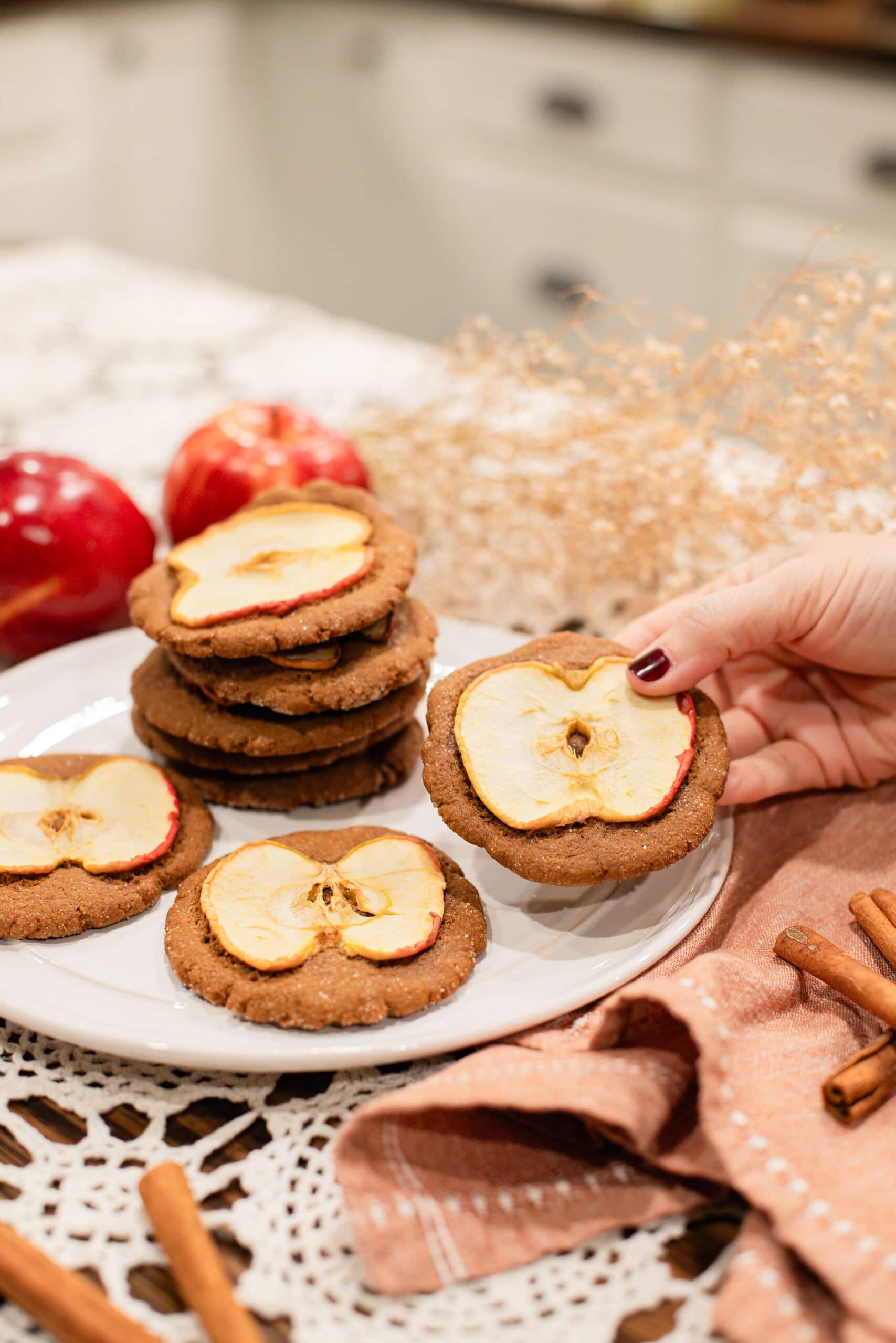 A hand holding a sourdough ginger apple snap cookie topped with a dried apple slice, with more cookies stacked on a white plate behind it, styled with red apples, cinnamon sticks, and dried botanicals on a lace doily.