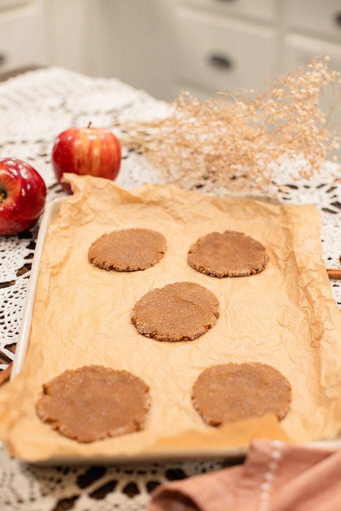 Five flattened sourdough ginger apple snap cookie rounds coated in cinnamon sugar arranged on a parchment-lined baking sheet before baking, styled with red apples and dried botanicals in a cottage kitchen.