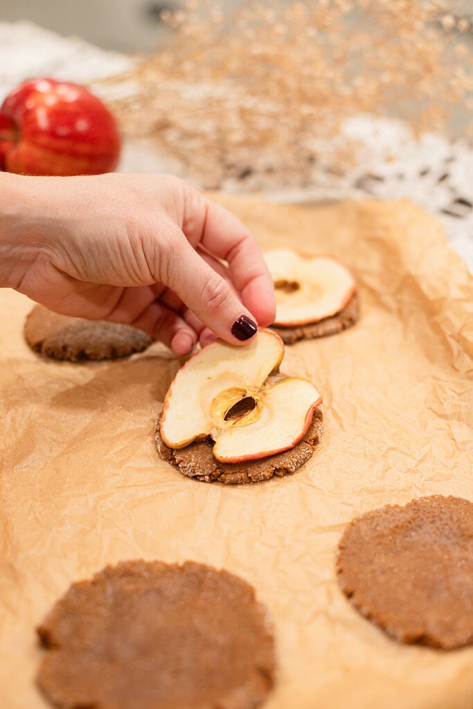 A hand placing a dried apple slice on top of an unbaked sourdough ginger snap cookie on a parchment-lined baking sheet, with more cookies visible around it and red apples softly blurred in the background.
