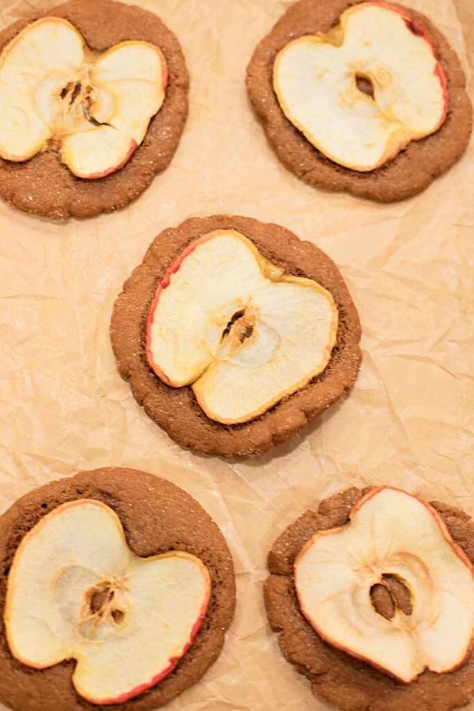 Overhead close-up of five baked sourdough ginger apple snap cookies each topped with a dried apple slice on a parchment-lined baking sheet.