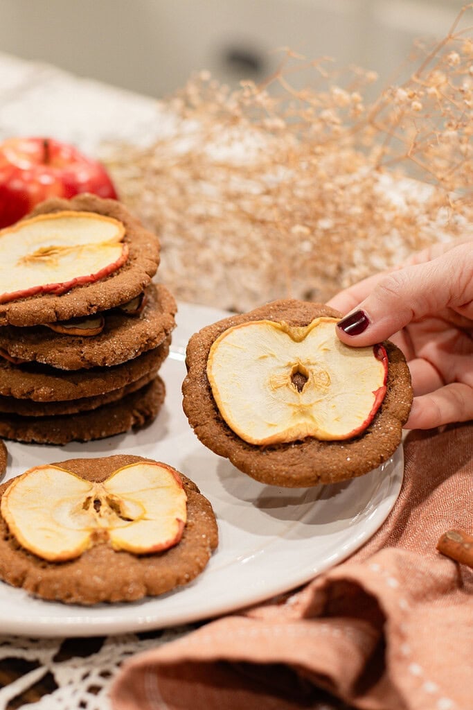A hand holding a sourdough ginger apple snap cookie topped with a dried apple slice over a white plate stacked with more cookies, styled with red apples, cinnamon sticks, dried botanicals, and a dusty rose linen.
