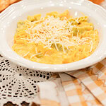 A white bowl of pumpkin mac and cheese with mixed yellow and green pasta in a golden pumpkin cheese sauce, topped with freshly grated parmesan, styled with two small orange pumpkins, a toasted roll, and dried baby's breath on a gold gingham linen.