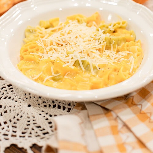 A white bowl of pumpkin mac and cheese with mixed yellow and green pasta in a golden pumpkin cheese sauce, topped with freshly grated parmesan, styled with two small orange pumpkins, a toasted roll, and dried baby's breath on a gold gingham linen.