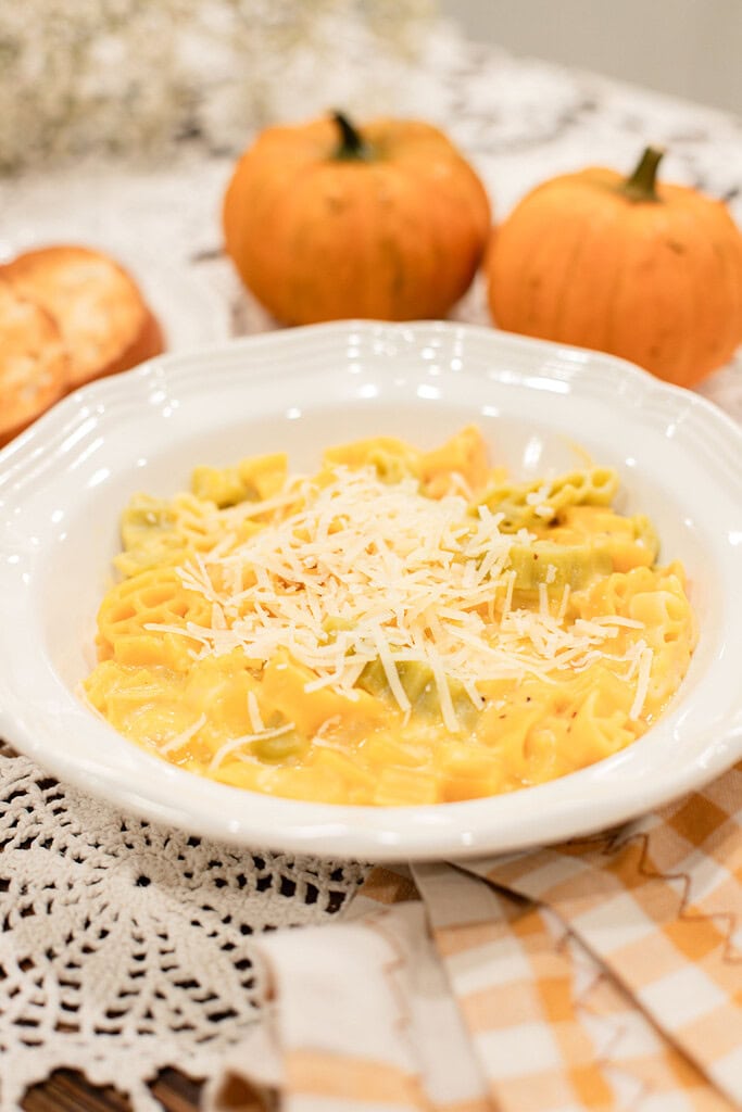 A white bowl of pumpkin mac and cheese with mixed yellow and green pasta in a golden pumpkin cheese sauce, topped with freshly grated parmesan, styled with two small orange pumpkins, a toasted roll, and dried baby's breath on a gold gingham linen.