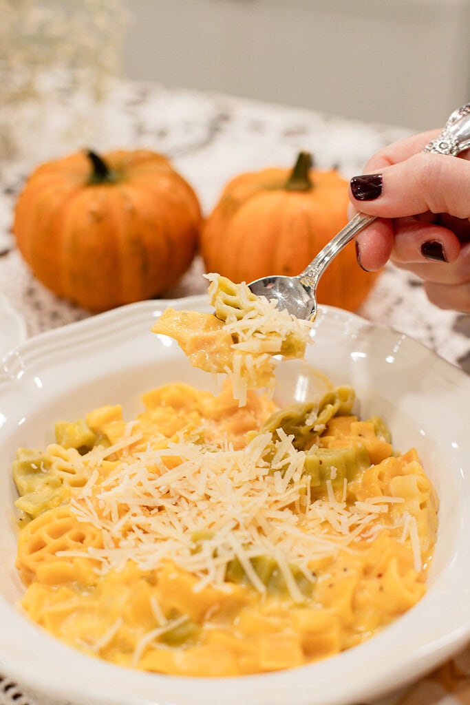 A hand lifting a spoonful of pumpkin mac and cheese from a white bowl, showing the creamy golden pumpkin sauce coating the mixed pasta and grated parmesan, with small orange pumpkins softly blurred in the background.