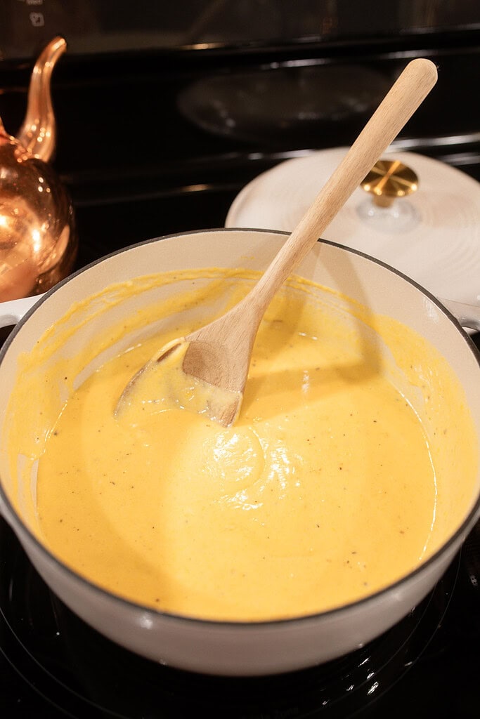 A white enameled Dutch oven filled with smooth golden pumpkin cheese sauce being stirred with a wooden spoon on the stovetop, with a copper kettle glowing in the background.