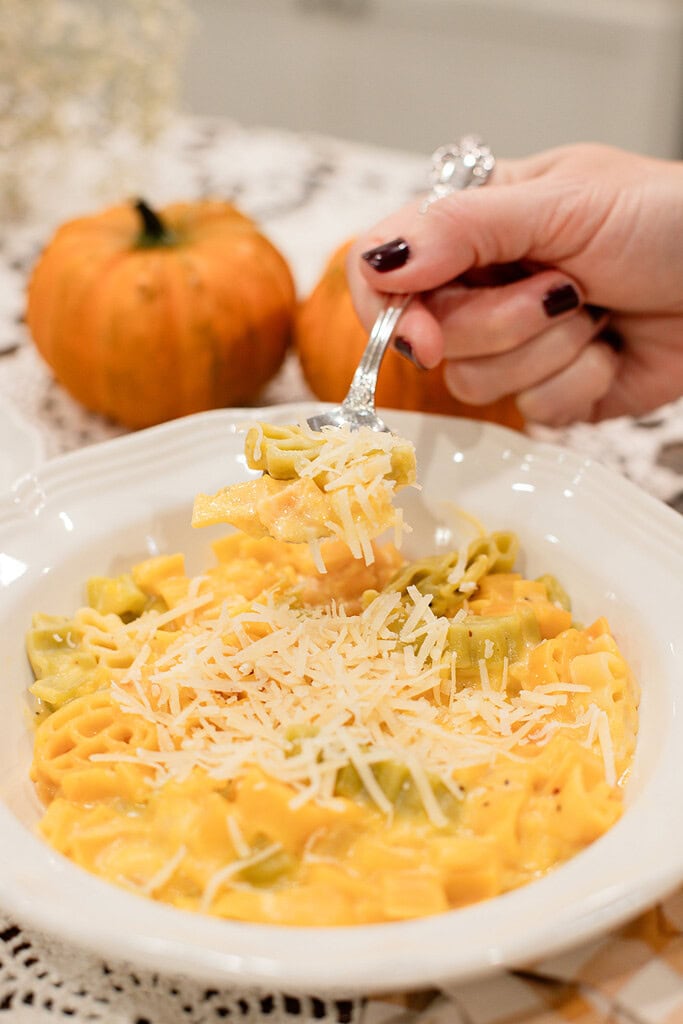 A hand holding a silver spoon lifting a creamy bite of pumpkin mac and cheese from a white bowl topped with grated parmesan, with orange pumpkins softly blurred behind it on a lace doily in a cottage kitchen.