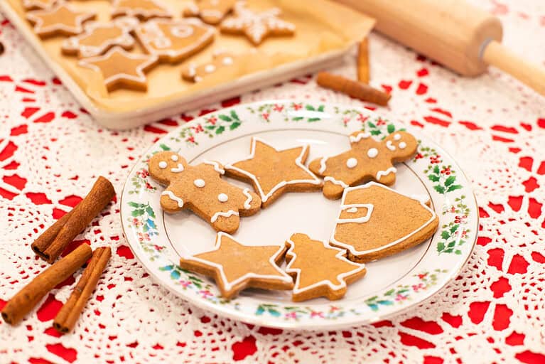 A festive plate of decorated sourdough gingerbread cookies featuring shapes like gingerbread men, stars, Christmas trees, and Santa hats outlined with white icing. The plate is set on a red and white lace-patterned surface with cinnamon sticks and a parchment-lined tray of additional cookies in the background. A rolling pin adds a cozy holiday baking touch.