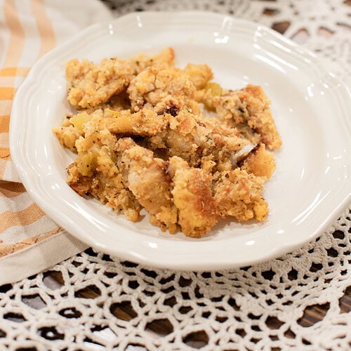 A close-up serving of sourdough cornbread dressing on a white plate, showing the golden crumbled cornbread and celery texture, with the baking dish softly blurred behind it on a gold gingham linen.
