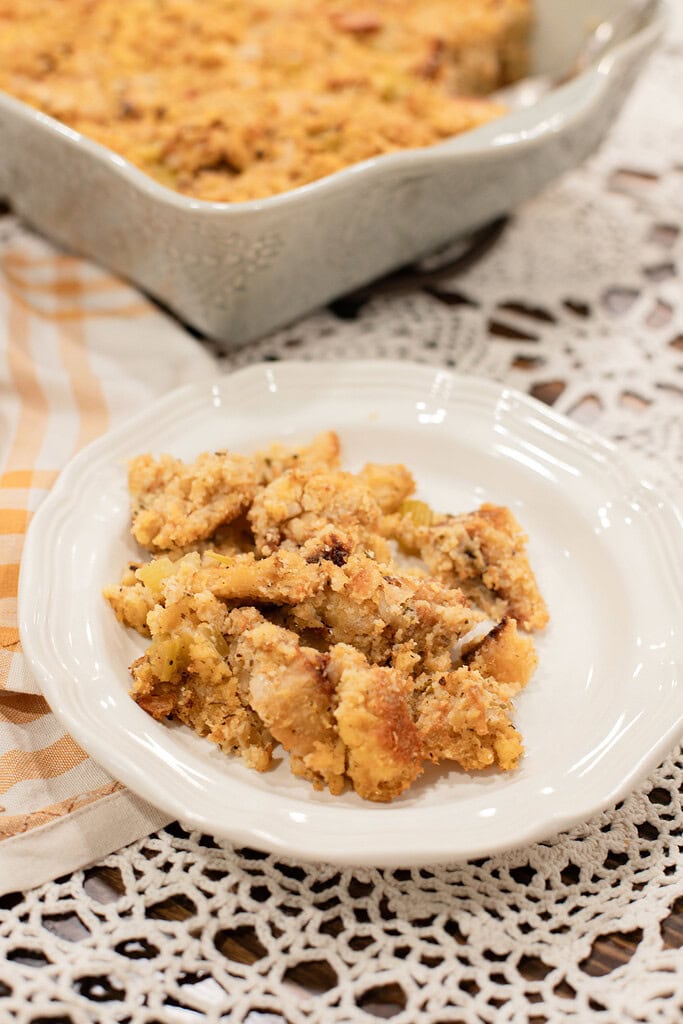 A close-up serving of sourdough cornbread dressing on a white plate, showing the golden crumbled cornbread and celery texture, with the baking dish softly blurred behind it on a gold gingham linen.