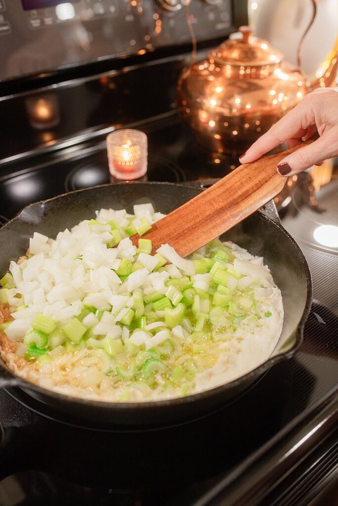 A hand stirring diced onion and celery sautéing in butter in a cast iron skillet on the stovetop, with a lit votive candle and a glowing copper kettle in the background.