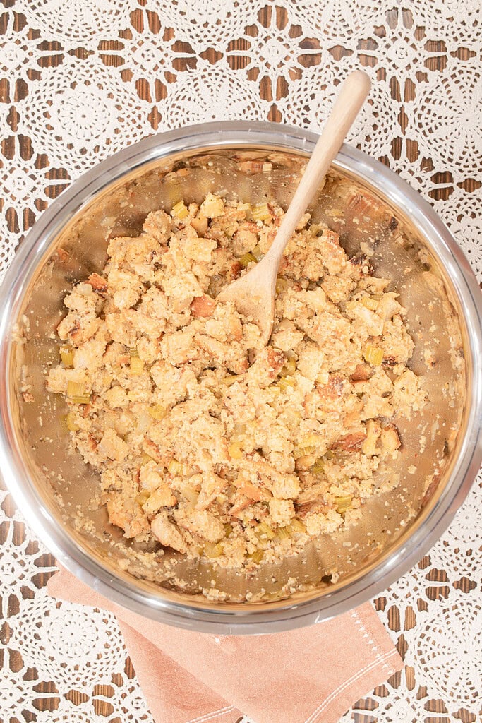 Overhead view of sourdough cornbread dressing mixture with crumbled cornbread, celery, and onion combined in a large stainless steel mixing bowl with a wooden spoon, on a lace doily with a dusty rose linen.