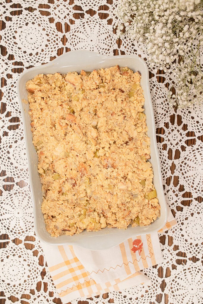 Overhead view of unbaked sourdough cornbread dressing spread in a white ceramic baking dish, showing the crumbled cornbread and celery mixture before going into the oven, styled on a lace doily with a gold gingham linen and dried baby's breath.