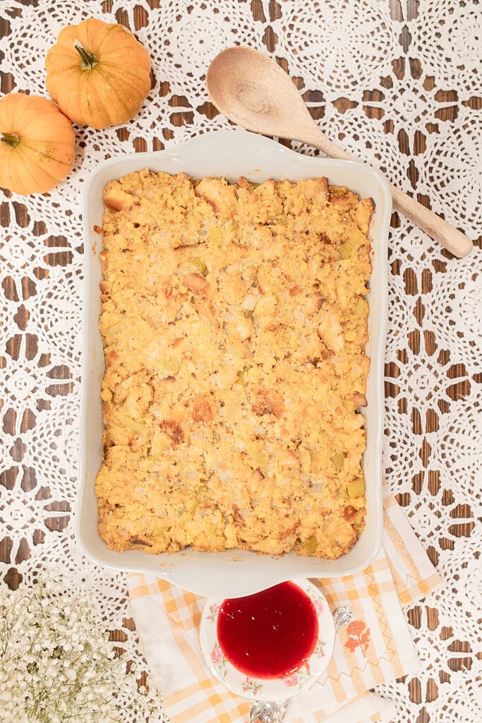 Overhead view of a fully baked sourdough cornbread dressing with a golden crust in a white ceramic baking dish, styled on a lace doily with mini pumpkins, a wooden spoon, a small dish of cranberry sauce, and a gold gingham linen.