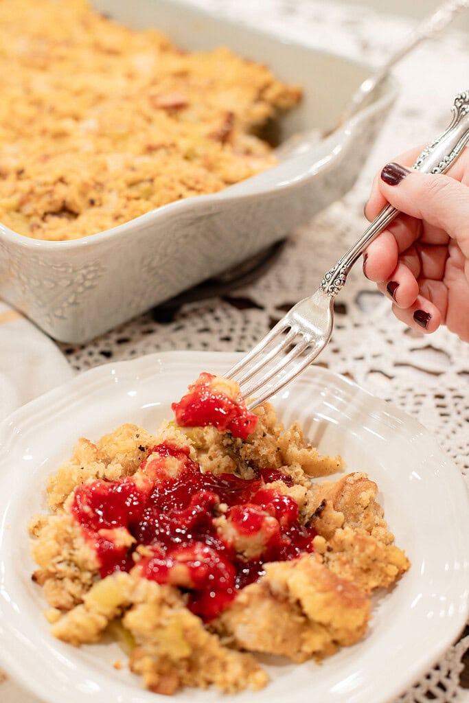 A hand holding a silver fork lifting a bite of sourdough cornbread dressing topped with bright red cranberry orange sauce from a white plate, with the full baking dish softly blurred behind it on a lace doily.