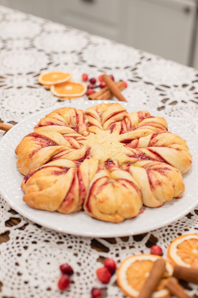 A golden baked sourdough cranberry orange star bread with crimson cranberry filling peeking through the twisted points on a white plate, styled on a lace doily with dried orange slices, fresh cranberries, and cinnamon sticks.