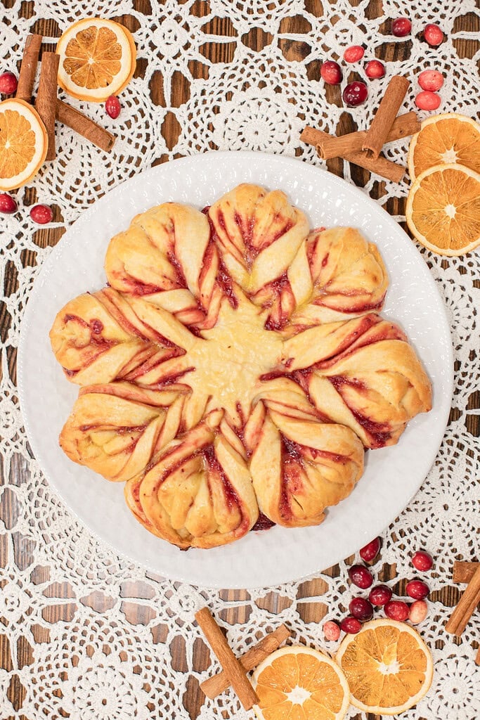 Overhead view of a sourdough cranberry orange star bread with golden twisted points and ribbons of cranberry filling on a white plate, styled on a lace doily with dried orange slices, fresh cranberries, and cinnamon sticks.
