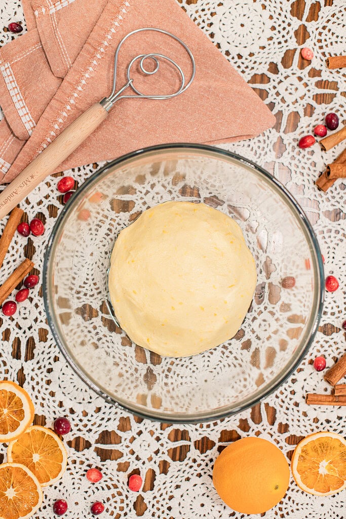 Overhead view of a smooth orange-zested sourdough dough ball in a glass bowl ready to rise, with a Danish dough whisk, a dusty rose linen, fresh cranberries, dried orange slices, and cinnamon sticks on a lace doily.