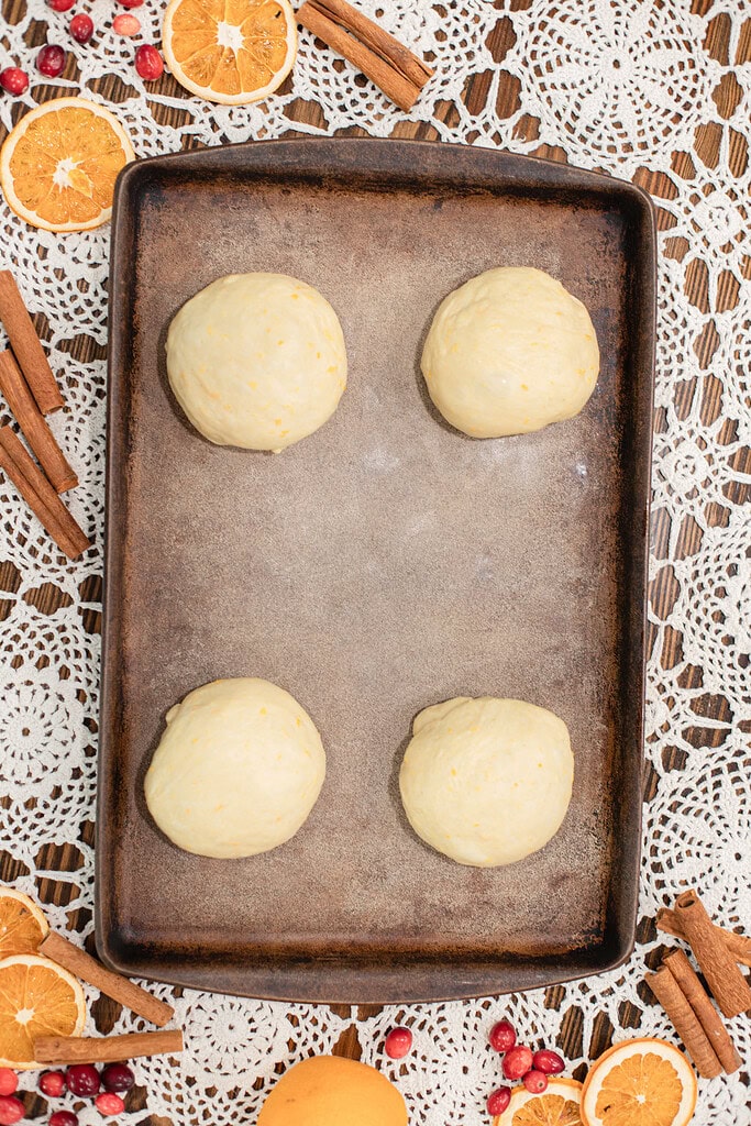 Overhead view of four smooth sourdough dough balls portioned on a parchment-lined dark baking sheet before shaping, styled on a lace doily with dried orange slices, cinnamon sticks, and fresh cranberries.