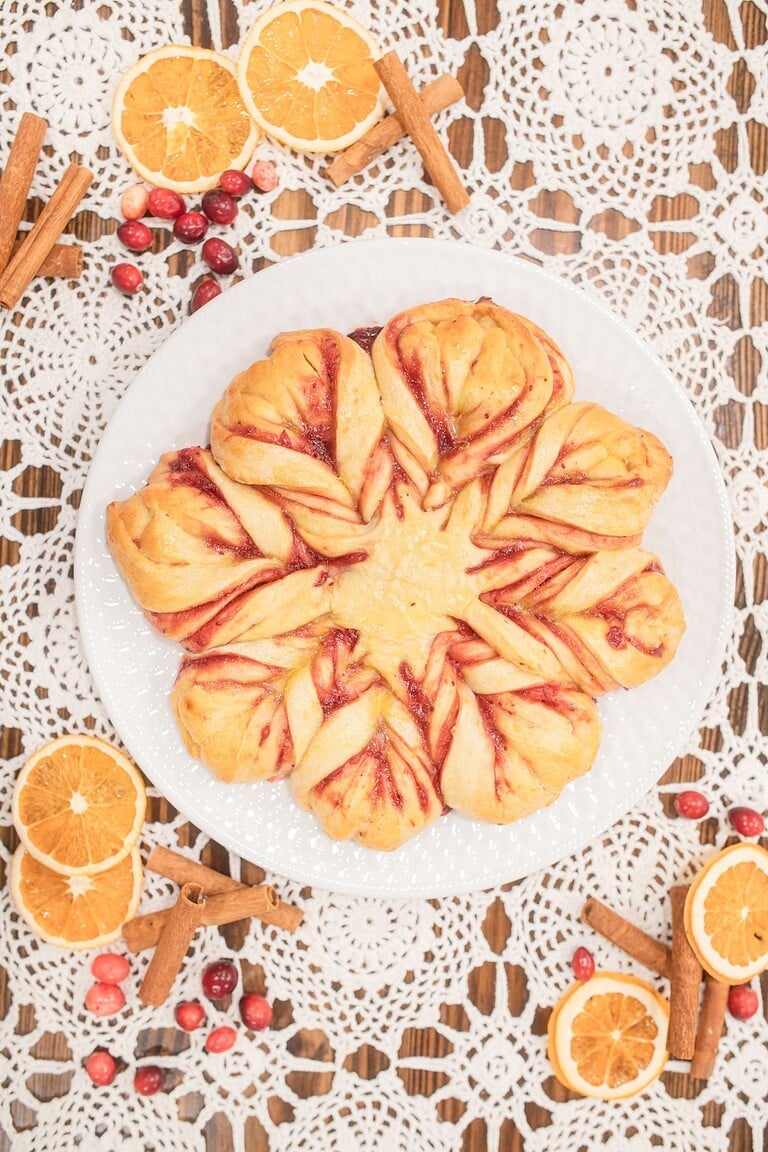 Overhead view of a freshly baked sourdough cranberry orange star bread on a white plate showing the golden crust with bright cranberry filling swirled through the twisted star points, on a lace doily with dried oranges and fresh cranberries.
